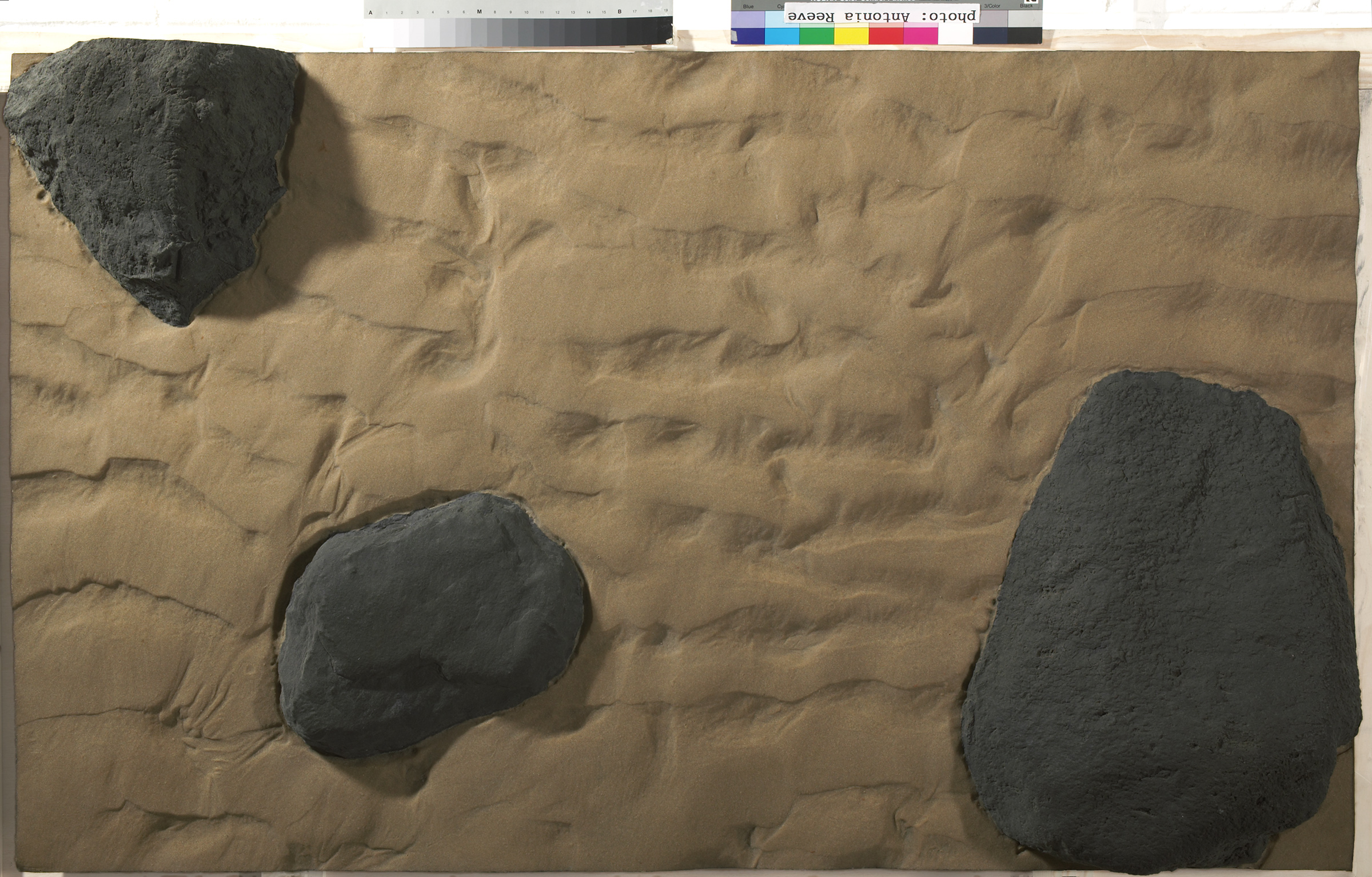 Fibreglass cast of a segment of sand and rocks, painted in realistic colours. Three black rocks protrude from rippled golden colour sand. 