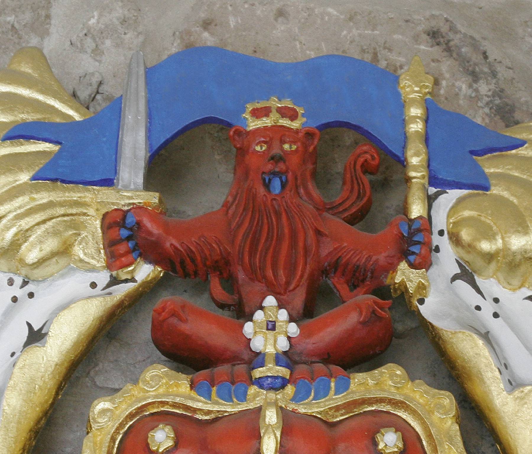 Two unicorns, each holding a Scottish flag, with a lion rampant on a shield in the middle. Unicorn on left holds the lion rampant flag. Unicorn on right holds the St. Andrews or saltire flag. At the top is a small lion griffin sitting on a crown. At the bottom is a small St. Andrew with his cross.