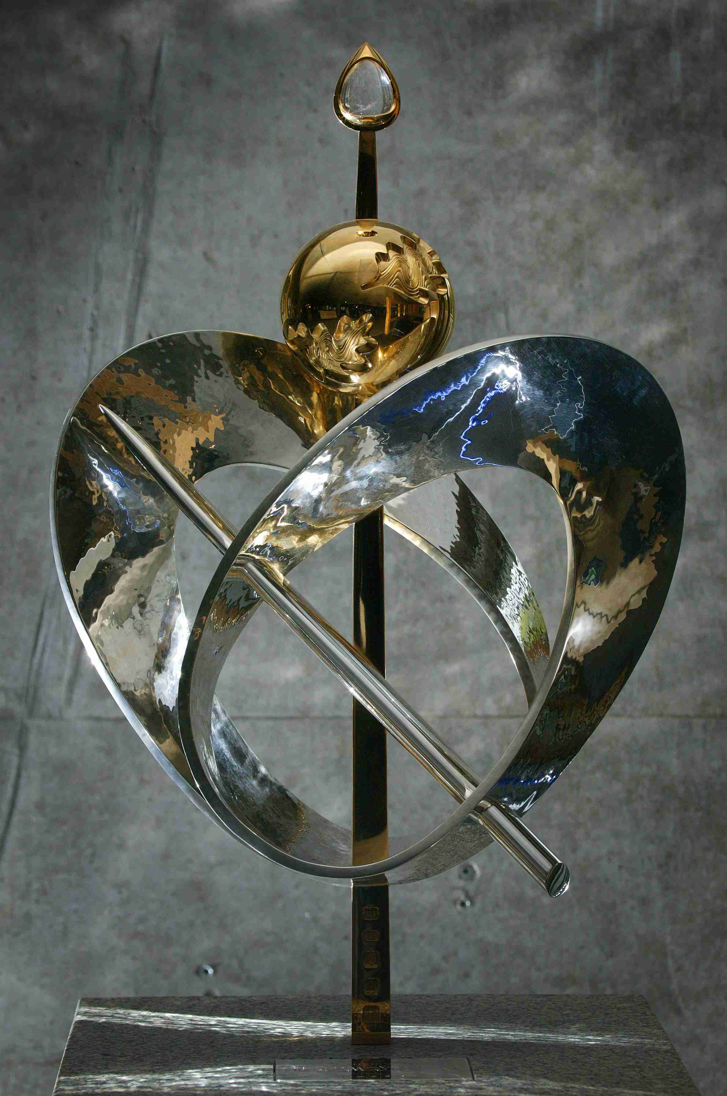 Abstract heart-shaped silver sculpture which is hollow in the centre. Through the centre is a gold staff with a gold ball on top, topped again by a rock crystal encased in gold. The heart shape is also intersected on the diagonal by a narrow silver pointed cylinder. Sculpture is inset into a solid granite plinth.